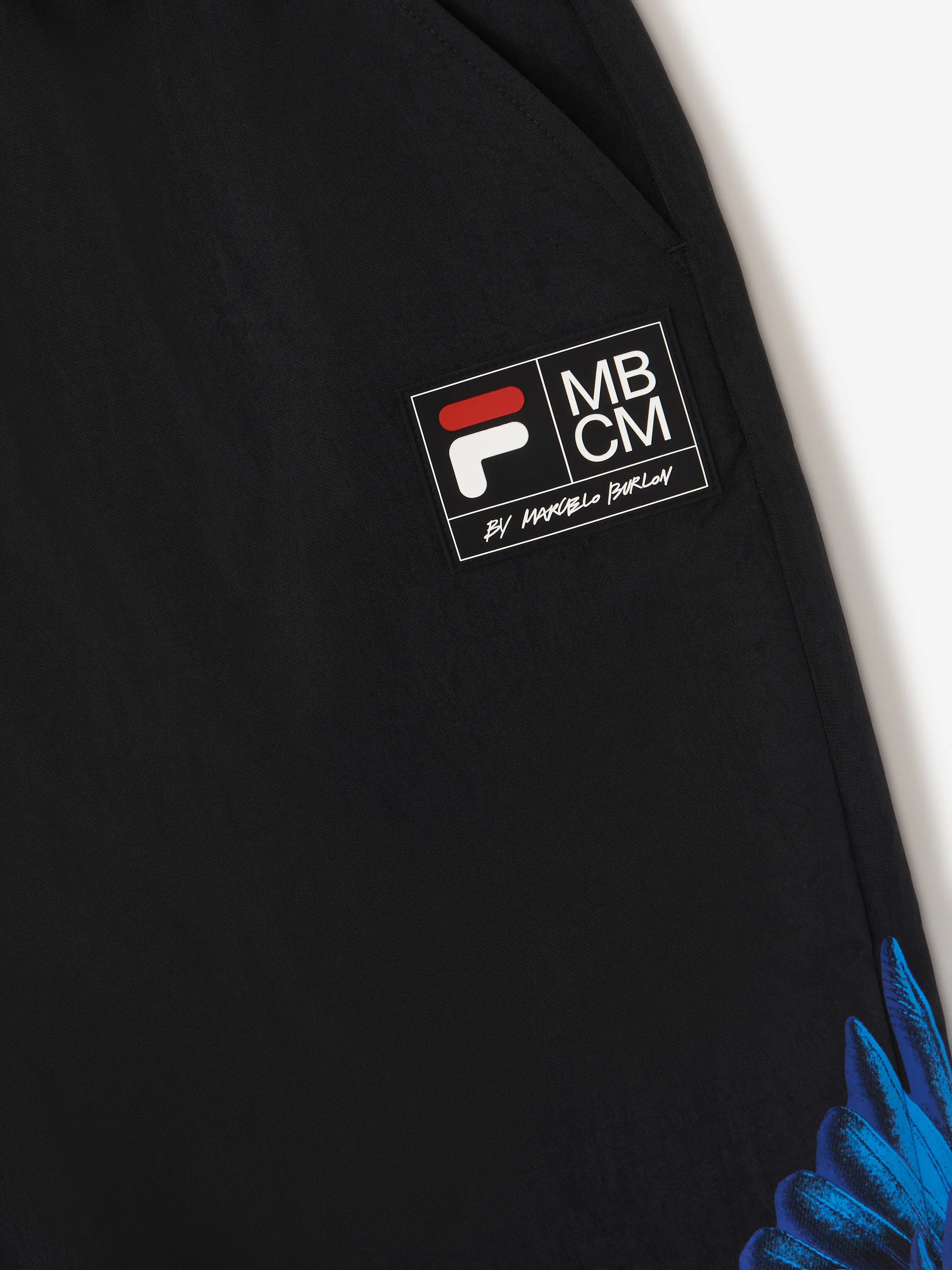 Black/blue x FILA feather print trousers from Marcelo Burlon County of Milan featuring feather print, logo patch to the side, slip pockets to the sides, elasticated waistband, front button and zip fastening and elasticated ankles.