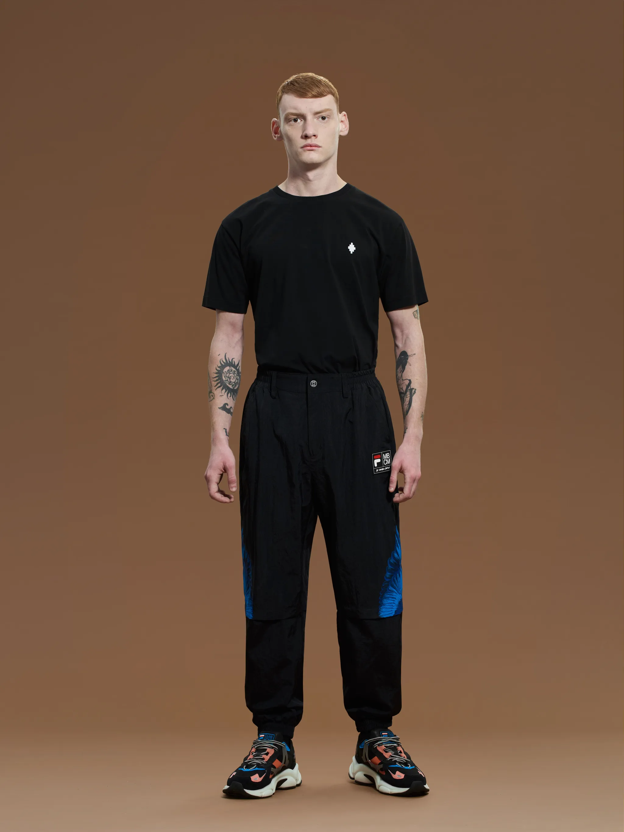 Black/blue x FILA feather print trousers from Marcelo Burlon County of Milan featuring feather print, logo patch to the side, slip pockets to the sides, elasticated waistband, front button and zip fastening and elasticated ankles.